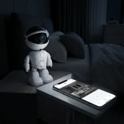 Бяла смарт камера FipTec с форма на робот е поставена на нощно шкафче в спалня. Смартфон до нея показва на живо видео в реално време, осигурявайки дискретна сигурност и лесен контрол за вашето пълно спокойствие.