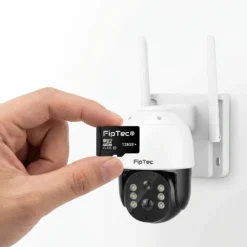 Ръка държи 128GB MicroSD карта FipTec пред бяла WiFi камера, монтирана на стена, осигурявайки надеждно локално съхранение на записите за пълна защита и спокойствие на вашия дом.