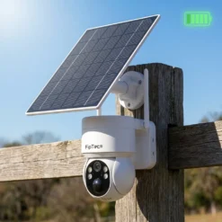 Бяла безжична соларна камера FipTec със соларен панел, монтирана на дървена ограда под ярко слънце, предлагаща автономна охрана и пълно спокойствие за вашия дом чрез зелена енергия.