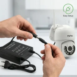 Ръце, свързващи захранващ кабел от 12V UPS батерия към бяла FipTec WiFi камера за видеонаблюдение върху бяла повърхност. Осигурете си лесен контрол и непрекъсната защита за пълно спокойствие у дома.