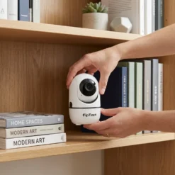Ръце, поставящи бяла вътрешна смарт WiFi камера FipTec на дървена етажерка с книги, показващи лесен монтаж и стилен дизайн. Осигурете си надеждна охрана и интелигентна сигурност за вашето пълно спокойствие чрез дискретно и лесно за позициониране видеонаблюдение в дома.
