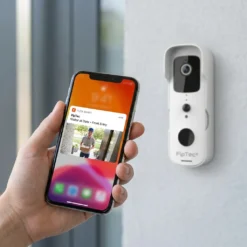 Ръка държи смартфон с известие за посетител от бял смарт видео звънец FipTec, монтиран на стена. Приложението показва реална картина на куриер пред входната врата, осигурявайки надеждна охрана и спокойствие за вашия дом чрез лесен контрол.