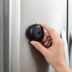 Женска ръка внимателно закрепва малка, черна, безжична WiFi камера FipTec на иноксовата врата на хладилник. Това е дискретно решение за сигурност, осигуряващо защита и спокойствие чрез лесна самоинсталация за вашия дом.