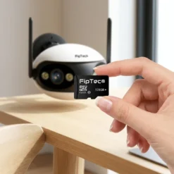 Ръка държи Micro SD карта памет FipTec 128GB пред панорамна WiFi камера LO18, осигурявайки надежден локален запис и сигурна защита на вашия дом за пълно спокойствие.