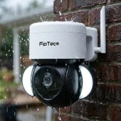 Бяла външна въртяща се смарт WiFi камера FipTec, демонстрираща своята водоустойчивост и надеждност при силен дъжд върху тухлена стена. Осигурете си надеждна охрана и интелигентна сигурност за вашето пълно спокойствие при всякакви метеорологични условия.