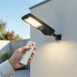 Ръка с дистанционно управление насочена към соларна LED лампа FipTec, монтирана на бяла външна стена. Осигурява интелигентна охрана и безжична сигурност за вашия дом и пълно спокойствие.