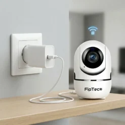 Бяла вътрешна Wi-Fi камера FipTec, поставена на дървена полица и включена в захранването. Сигурна безжична връзка, гарантираща пълна защита на дома и дискретно спокойствие за вашето семейство.