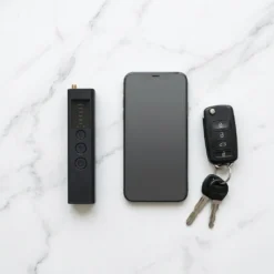 Детектор FipTec XScan LM10 е разположен до смартфон и ключове върху мраморен плот, демонстрирайки своя компактен и преносим размер. Уредът предлага дискретна защита и надеждна сигурност за вашето спокойствие.